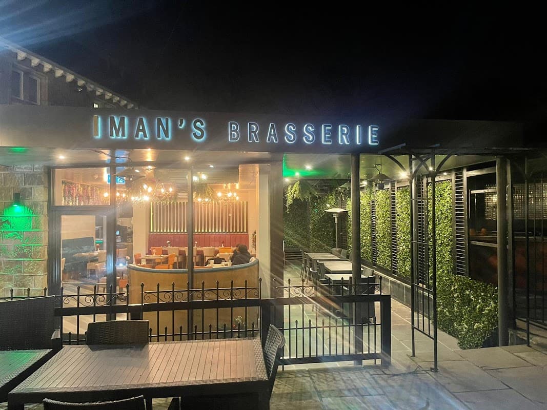 "Iman's Brasserie sign illuminated at night, with outdoor seating and green foliage in a cozy setting."