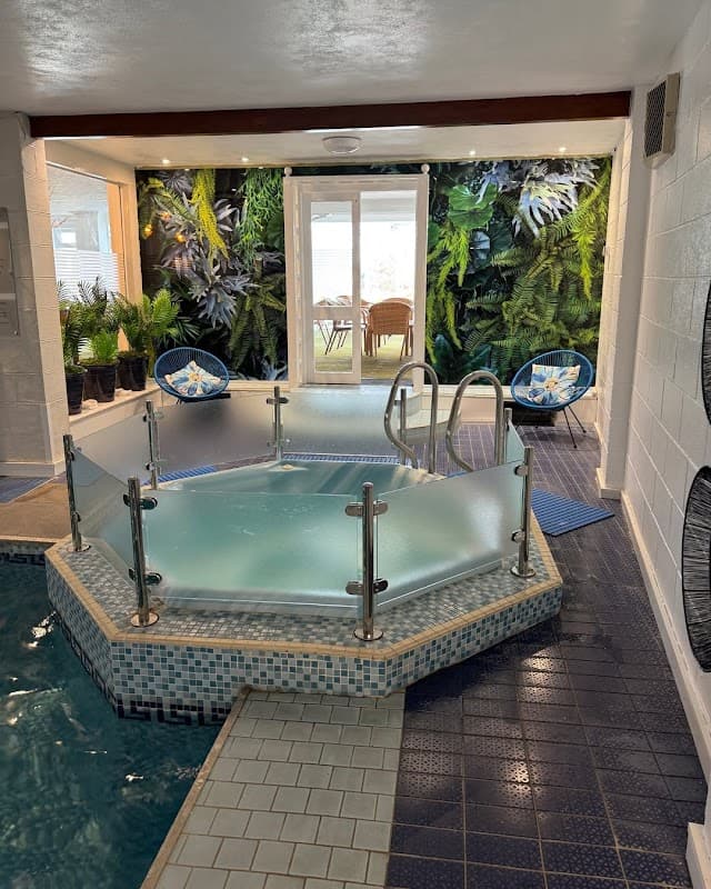 Hexagonal hot tub surrounded by lush greenery, with blue chairs and a doorway leading to a bright outdoor area.