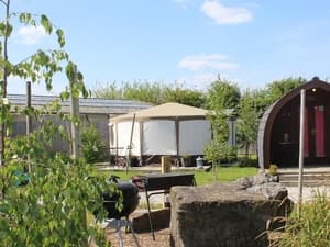 Curved wooden camping pod and white yurt in a grassy area with trees and rocks under a clear blue sky.